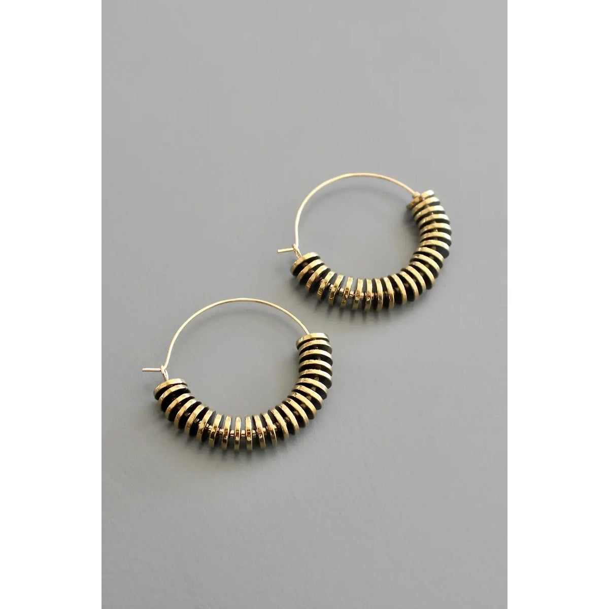 The DAVID AUBREY - GOLD HEMATITE HOOP EARRINGS feature gold-plated hoops embellished with stacked black and gold circular beads, artfully presented against a gray background. These DAVID AUBREY earrings bring a modern edge to any ensemble.