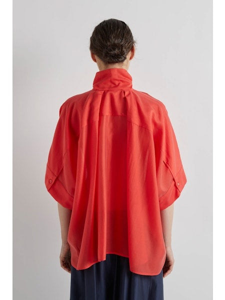 A person in profile wears the MEIMEIJ - SCARF TIE NECK BLOUSE by MEIMEIJ in bright red with short sleeves, paired with a dark, flowing skirt. Their dark hair is tied back, hands in pockets, against a plain white background.