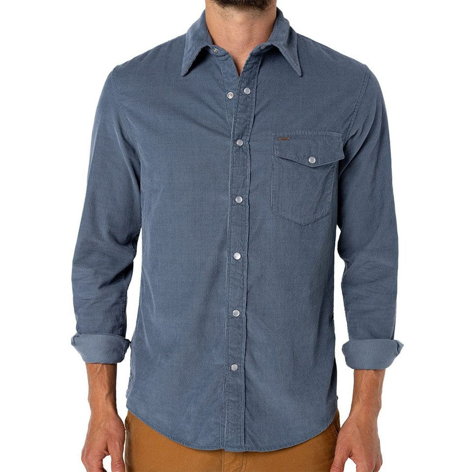 A man is wearing the CRIQUET Long Sleeve Corduroy Pearl Snap Shirt in Slate Blue by CRIQUET, featuring a chest pocket and rolled-up cuffs. He pairs it with brown pants against a white background.