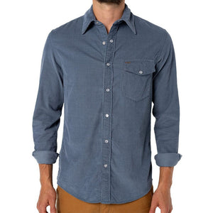 A man is wearing the CRIQUET Long Sleeve Corduroy Pearl Snap Shirt in Slate Blue by CRIQUET, featuring a chest pocket and rolled-up cuffs. He pairs it with brown pants against a white background.