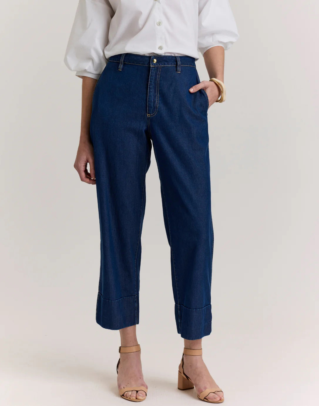 A person wearing a white button-up shirt, tan heeled sandals, and HINSON WU’s ROSA DARK WASH DENIM CROPPED PANT, standing against a plain light background.