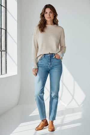 A woman stands against a white background in the KINROSS CASHMERE Doubleknit Lurex Trim Funnel Sweater in Dune and light blue jeans, her long hair pulled back as she smiles at the camera—an effortless travel look.