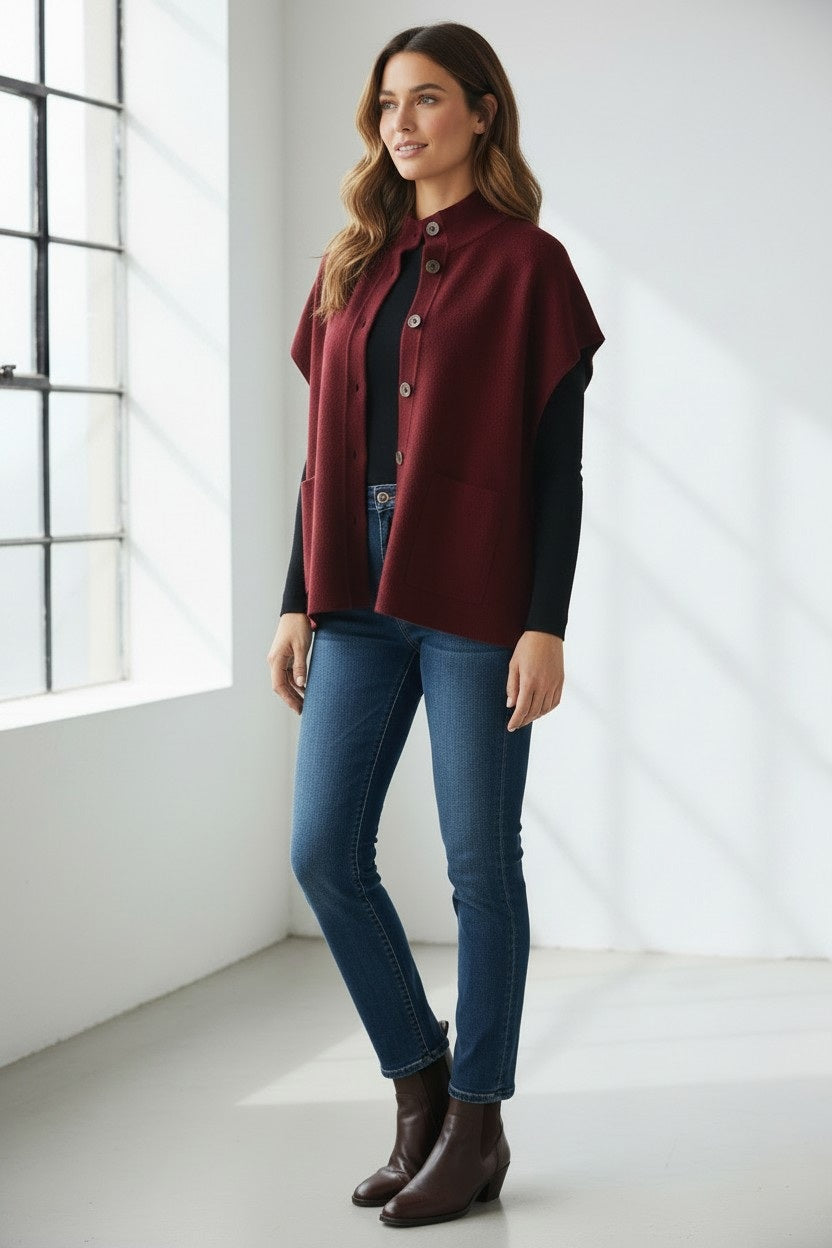 A woman stands indoors by a window, smiling softly as she wears the KINROSS CASHMERE Short Sleeve Button Mock Cardigan in a rich burgundy hue over a black top, blue jeans, and brown ankle boots. Her long hair falls freely around her shoulders.