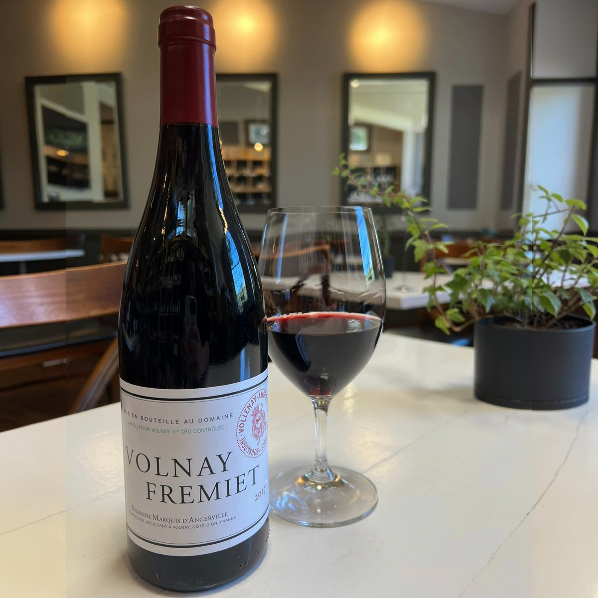 A bottle of MARQUIS D’ANGERVILLE VOLNAY 1ER CRU ‘FREMIET’ 2017 with a white label sits next to a glass of red wine on a white table.