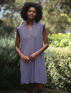 A person smiles outdoors, wearing the FRANK & EILEEN Mini Lily Popover Henley Tank Dress in Vintage Jersey Yacht Stripe, a sleeveless navy and white striped dress made in California. Lush greenery and a bright blue sky appear in the background.