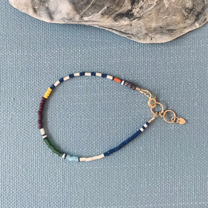 The ALICE RISE Thinnest Line Bracelet in Navy and Olive, featuring Japanese glass beads and gold clasps, is displayed on a blue textured surface with a large seashell partially visible.