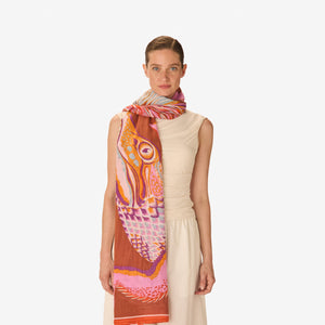 A woman wearing a light beige top and the INOUI EDITIONS - CROCO SCARF IN CARAMEL by INOUI, featuring vibrant pink, purple, red, and white feather-like patterns, draped over her shoulder.