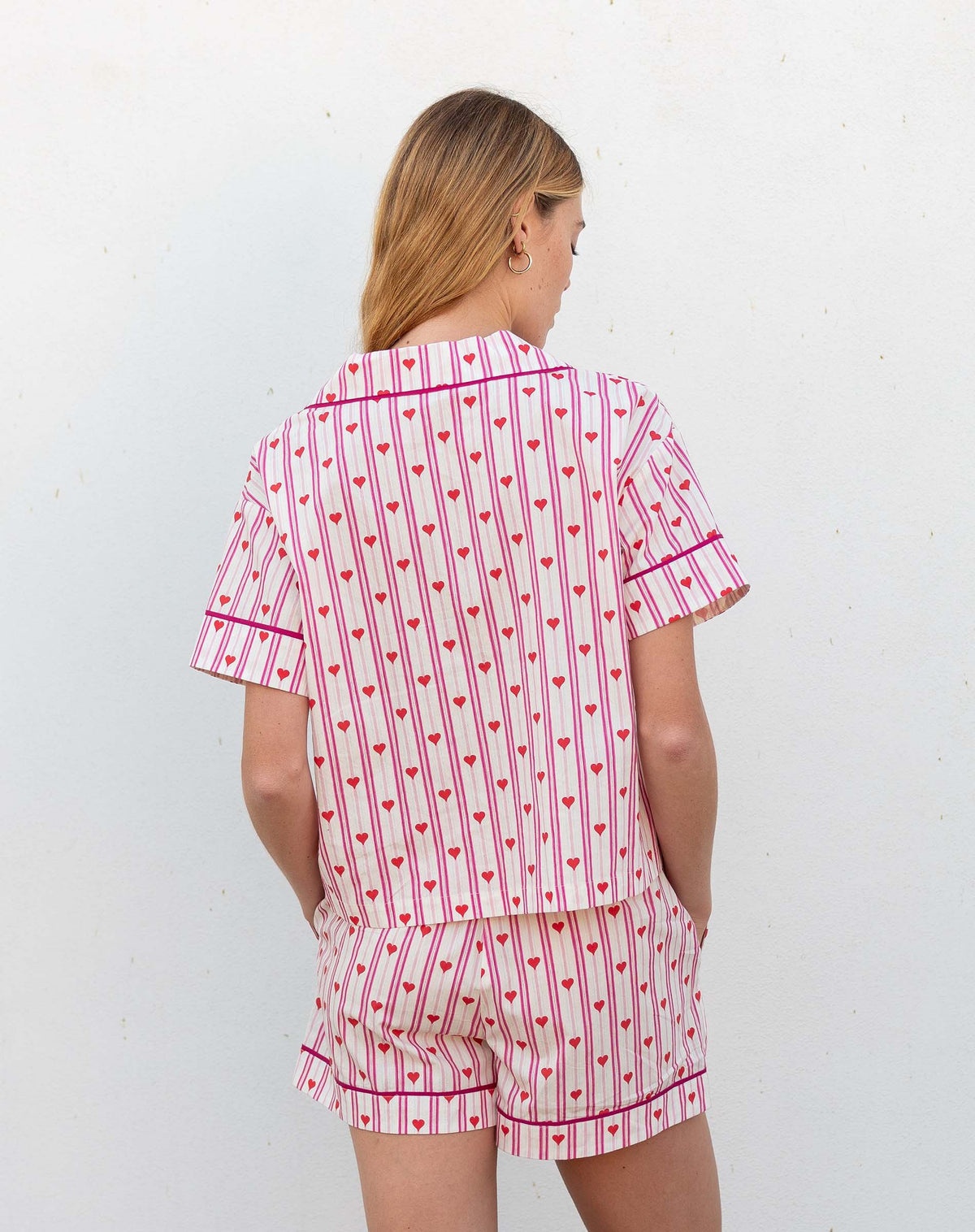 A woman with light brown hair smiles in MERSEA&#39;s OVER THE MOON PAJAMA SHORT SET LOVE NOTES— a heart print cotton short-sleeve button-up top and shorts—standing against a white background.