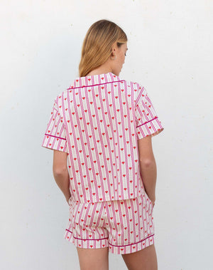 A woman with light brown hair smiles in MERSEA's OVER THE MOON PAJAMA SHORT SET LOVE NOTES— a heart print cotton short-sleeve button-up top and shorts—standing against a white background.