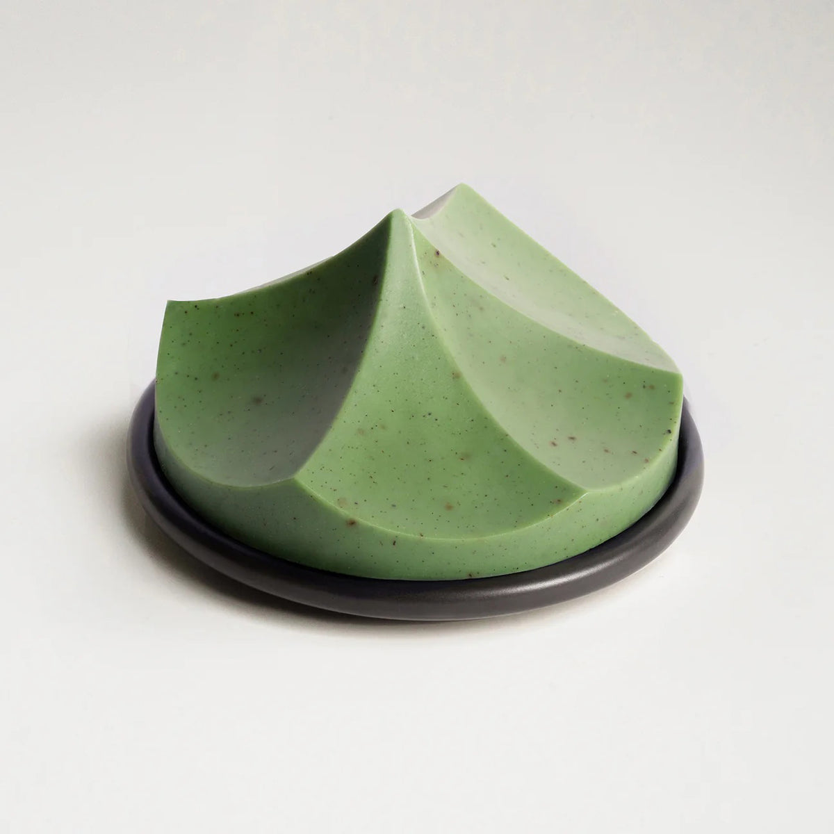 The UME – HERBES DE PROVENCE ERODE SOAP by UME STUDIO is a sculptural, geometric light green bar with subtle speckles and three peaked curves, displayed on a round dark soap dish against a plain white background.