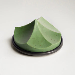The UME – HERBES DE PROVENCE ERODE SOAP by UME STUDIO is a sculptural, geometric light green bar with subtle speckles and three peaked curves, displayed on a round dark soap dish against a plain white background.