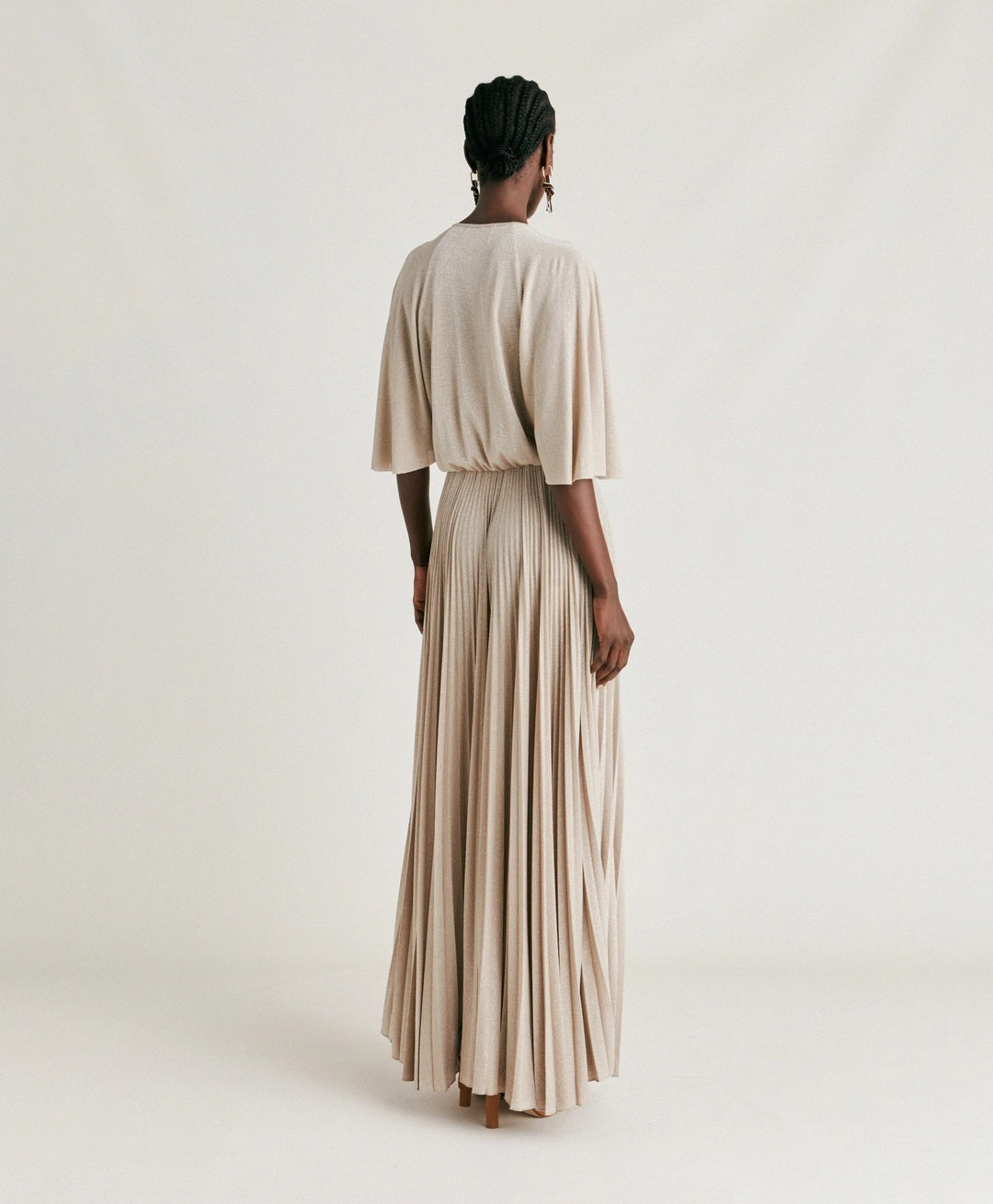 A person stands facing the camera, wearing the MOMONI - PARADISU DRESS, a light-colored, flowing pleated outfit with wide pants and kimono sleeves. Their hair is styled in neat braids, and they accessorize with drop earrings. The neutral background highlights the elegance of the MOMONI dress.