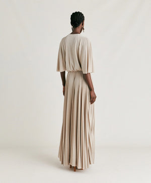 A person stands facing the camera, wearing the MOMONI - PARADISU DRESS, a light-colored, flowing pleated outfit with wide pants and kimono sleeves. Their hair is styled in neat braids, and they accessorize with drop earrings. The neutral background highlights the elegance of the MOMONI dress.