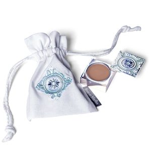 A small embroidered white drawstring pouch sits beside an open compact of ROSE ET MARIUS - A ROSE WINE UNDER THE ARBOR SOLID PERFUME, nestled in a decorative blue and white box, capturing the refined elegance of this Provençal fragrance.