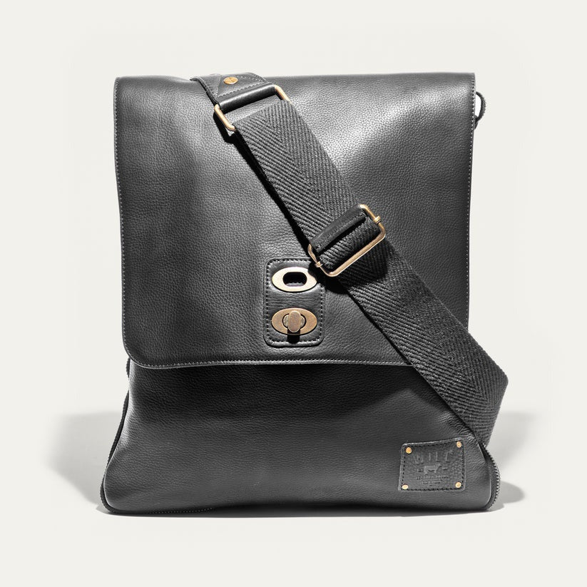 The WILL LEATHER GOODS OTTO Crossbody Bag in black full-grain leather features a front flap, gold-tone hardware, adjustable shoulder strap, and a small stitched patch on the lower right corner. Displayed against a plain white background.