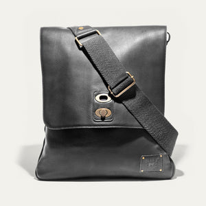 The WILL LEATHER GOODS OTTO Crossbody Bag in black full-grain leather features a front flap, gold-tone hardware, adjustable shoulder strap, and a small stitched patch on the lower right corner. Displayed against a plain white background.