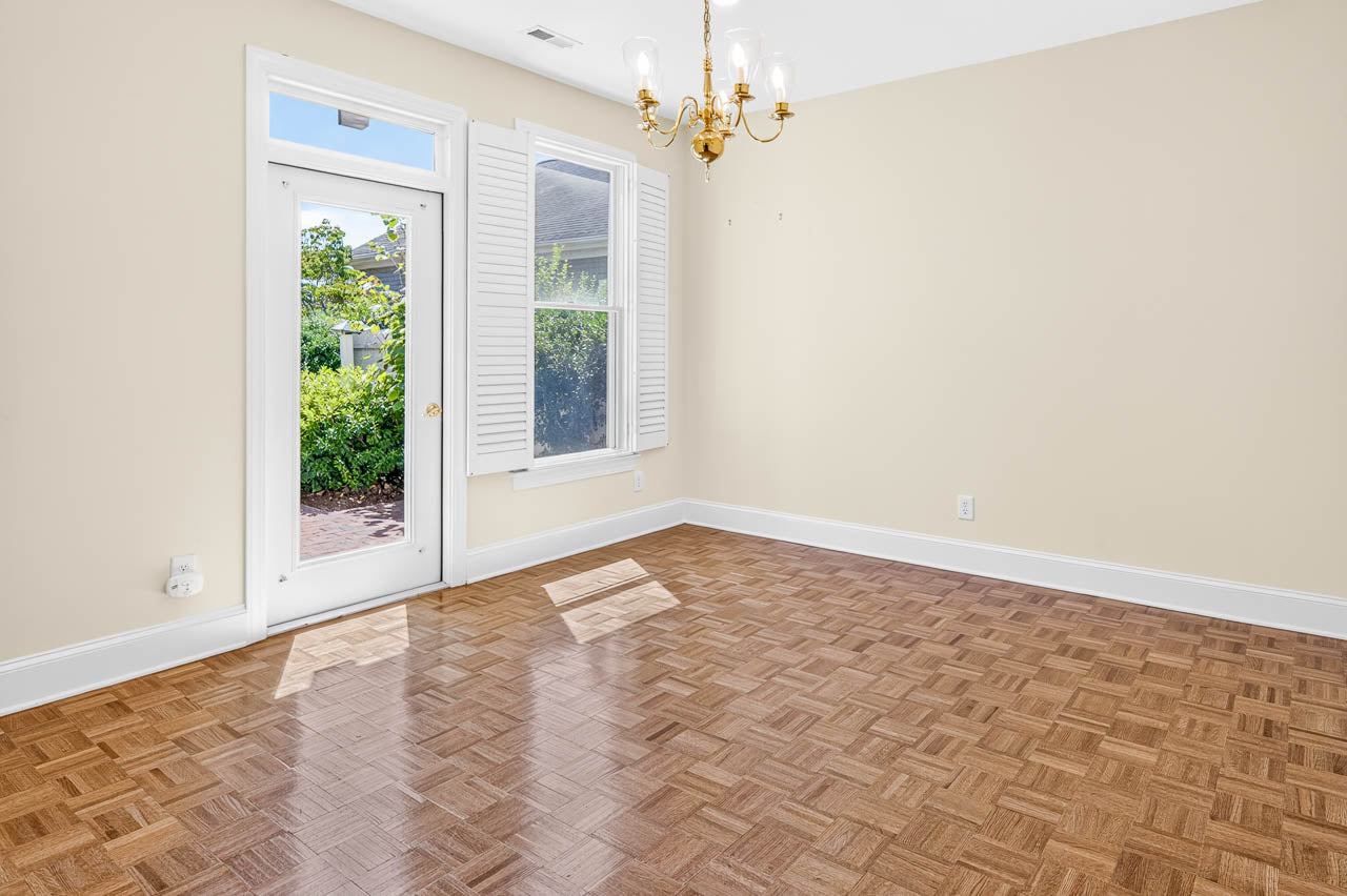Empty room with parquet wood floors, beige walls, a gold chandelier, white trim, one window with shutters, and a glass door leading outside to a patio with greenery visible.