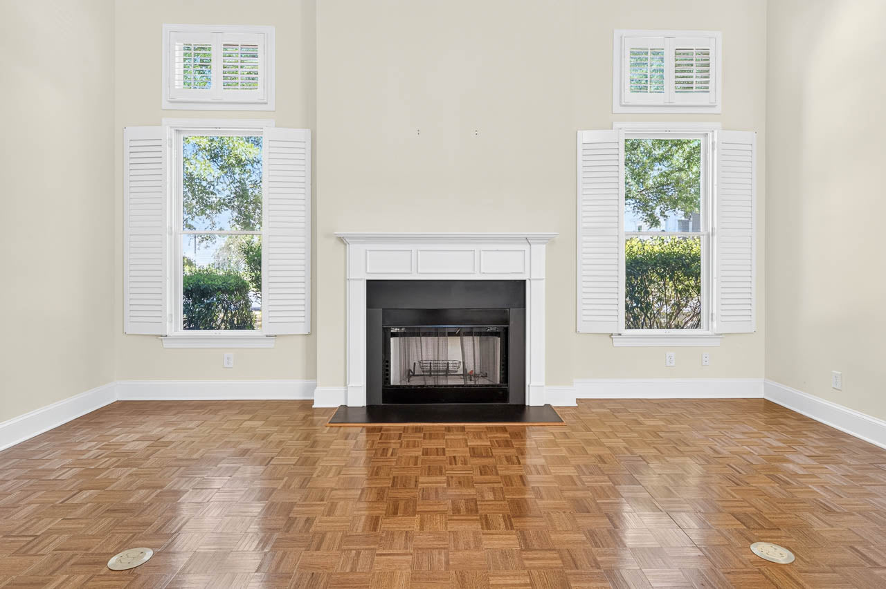 Bright living room with wooden parquet floor, white fireplace centered between two tall windows with open white shutters, and cream-colored walls.