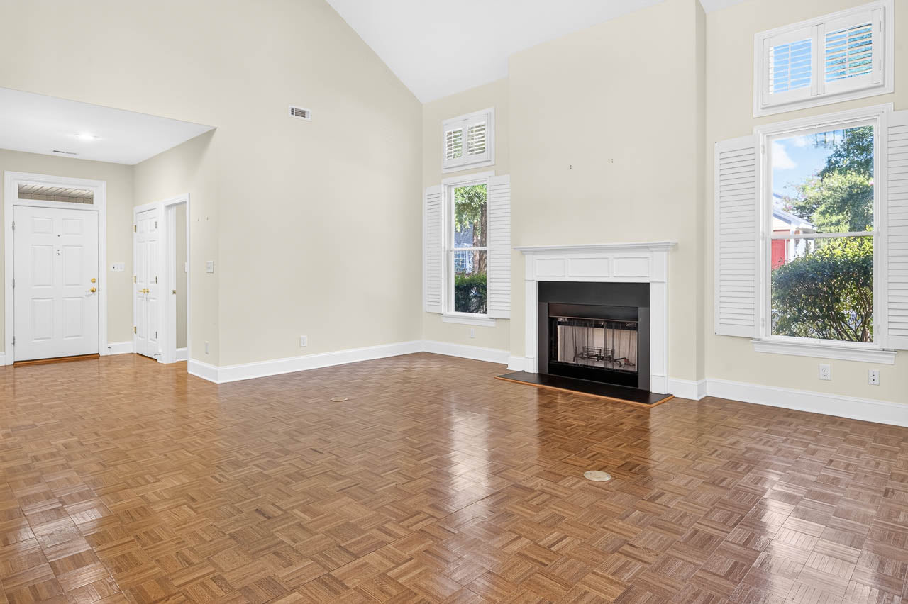 Spacious, empty living room with parquet wood floors, a white fireplace, tall ceilings, and large windows with shutters. Entryway and door are visible to the left. Neutral-colored walls create a bright, airy feel.