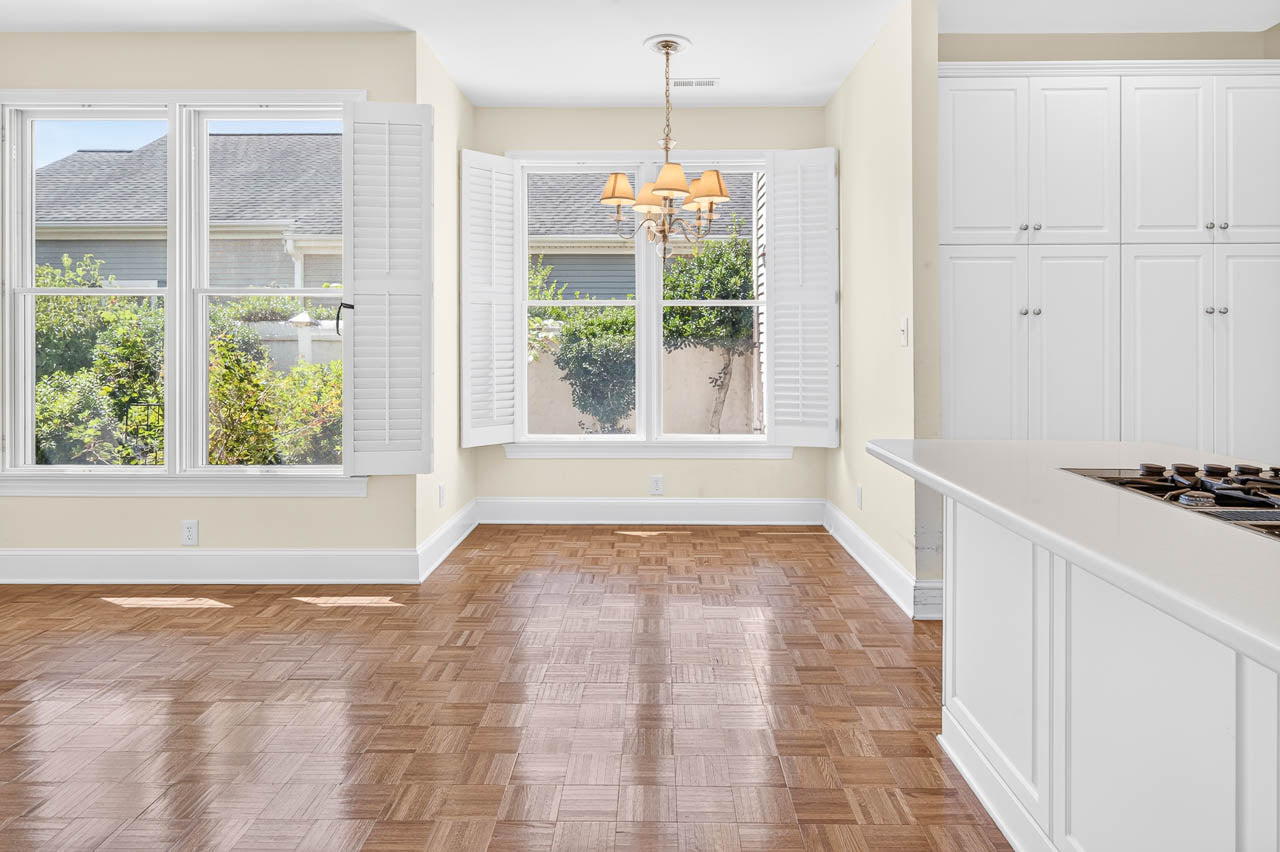 Bright, empty dining area with large windows, white shutters, wood parquet floor, chandelier, and a white kitchen island with a cooktop. Greenery is visible outside through the windows.