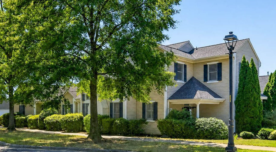 A two-story beige house with dark shutters and a gabled roof is surrounded by green trees and bushes. A black streetlamp stands near the sidewalk under a clear blue sky.