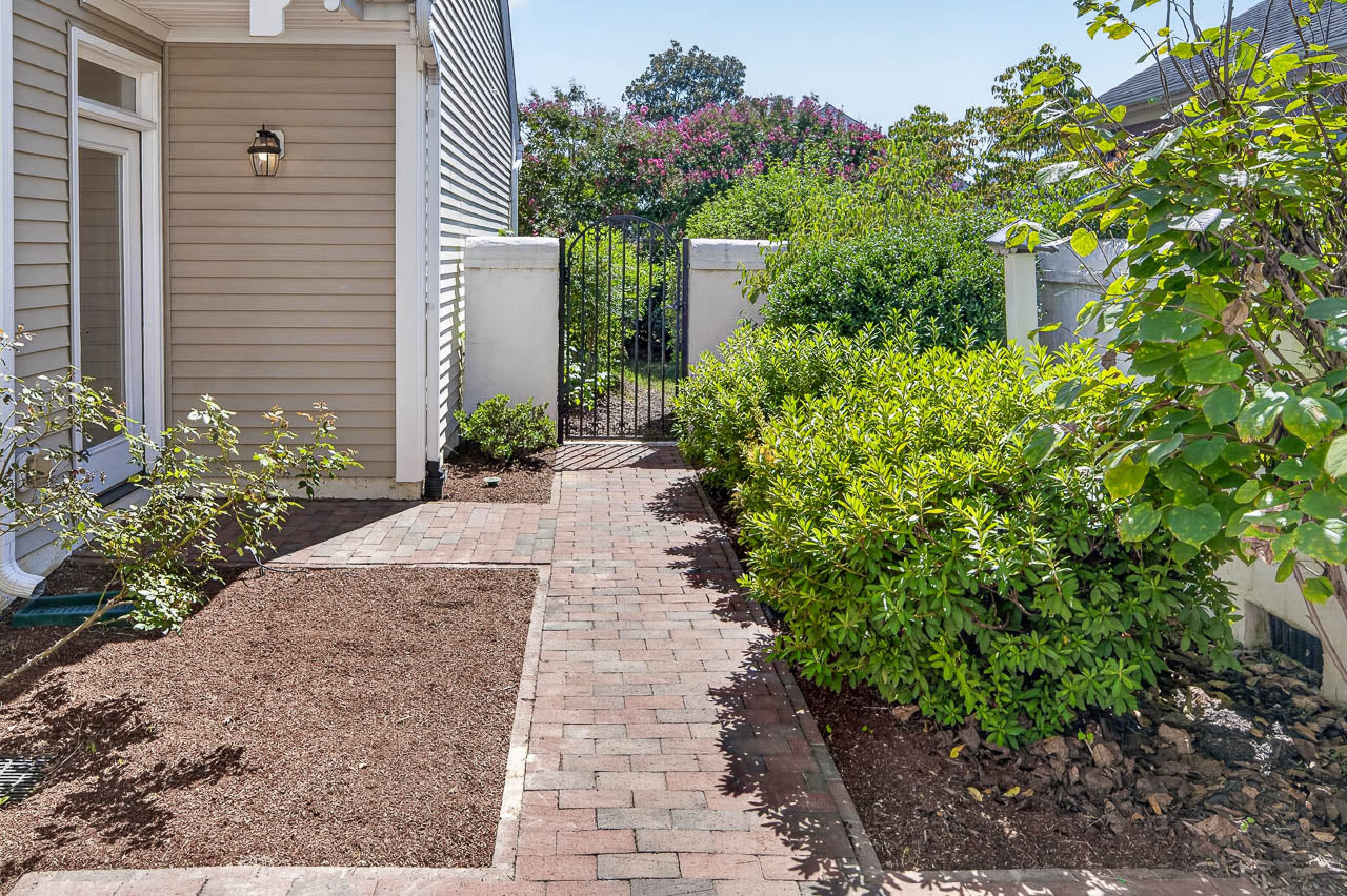 A brick walkway leads between green shrubs to a black metal gate, bordered by tan siding on the left and white walls in the background, with trees and plants beyond the gate.