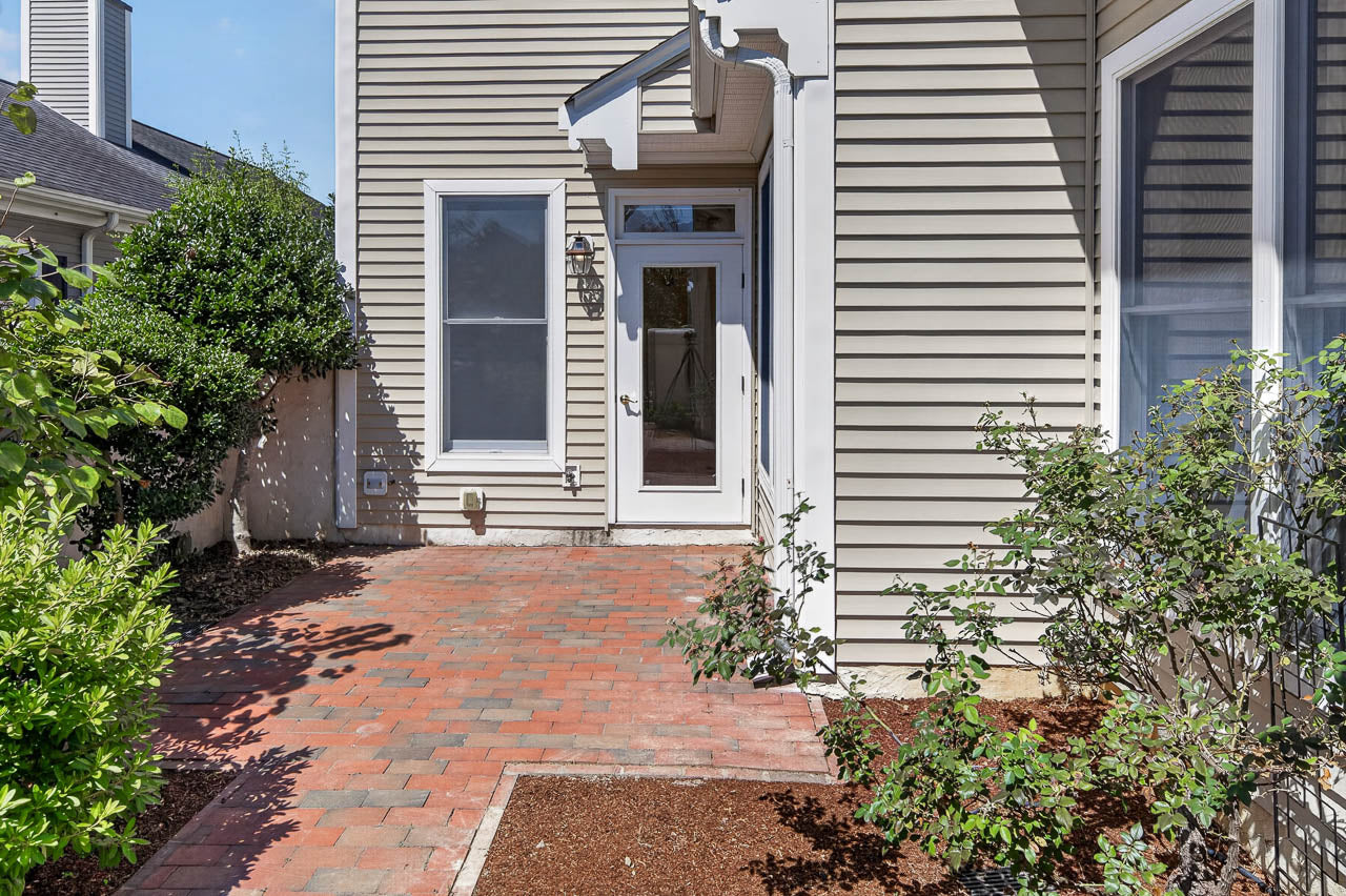 A brick patio with shrubs and bushes leads to a white door and window on a beige-sided house. Sunlight casts shadows on the ground and walls.