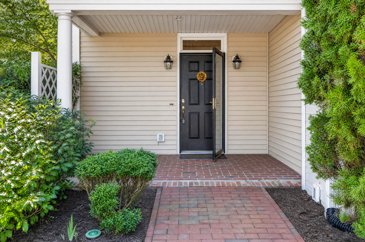 Open black front door with a wreath, two wall lanterns, beige siding, brick porch, and pathway surrounded by bushes and greenery.