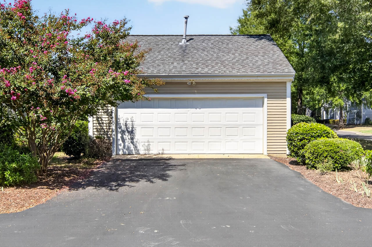 A two-car garage with a white door, beige siding, and a blacktop driveway, surrounded by green shrubs, a flowering tree, and mulch landscaping on a sunny day.