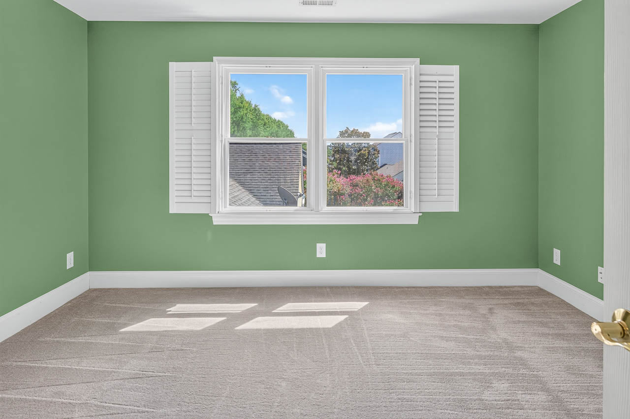 An empty room with light green walls, beige carpet, white window trim, and open white shutters. Sunlight streams through the window, revealing a view of rooftops, trees, and a blue sky with clouds.