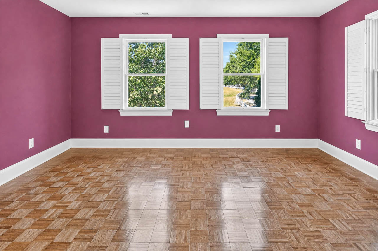 Empty room with purple walls, white trim, parquet wood floor, two windows with white shutters, and sunlight shining in, revealing a view of green trees outside.