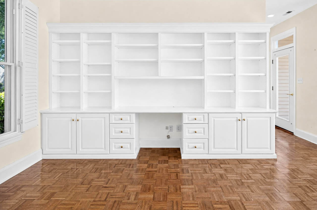 White built-in shelves and cabinets with gold handles are set against a cream wall on a parquet wood floor, with a large window on the left and a door on the right. The shelves are empty.