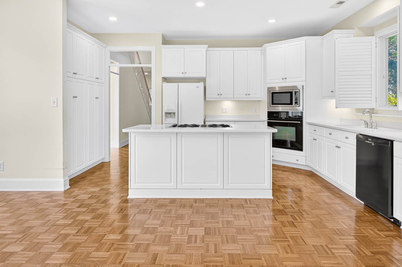 Modern kitchen with white cabinets, island with stove, built-in oven, microwave, dishwasher, large fridge, and parquet wood flooring. Bright space with natural light from a window above the sink.