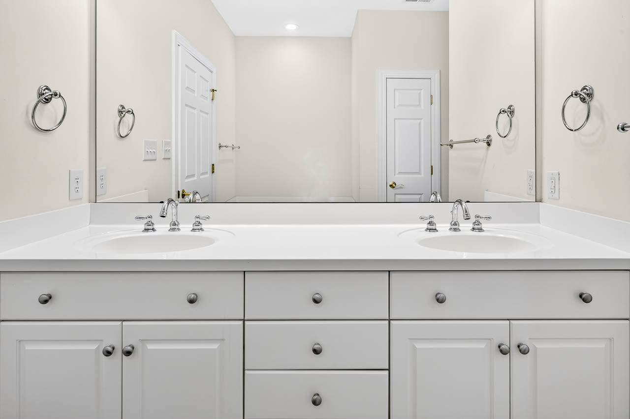 A modern bathroom features a double vanity with white cabinets, chrome faucets, and large mirrors reflecting the room. Two towel rings are mounted on either side of the sinks.