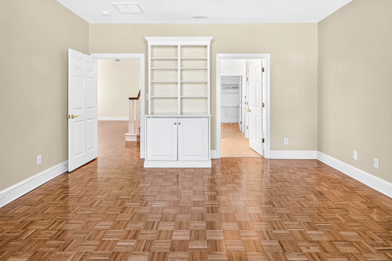 Room with parquet floor, beige walls, white built-in shelves with cabinets, two open doors leading to adjoining rooms, and visible staircase and closet areas in the background.