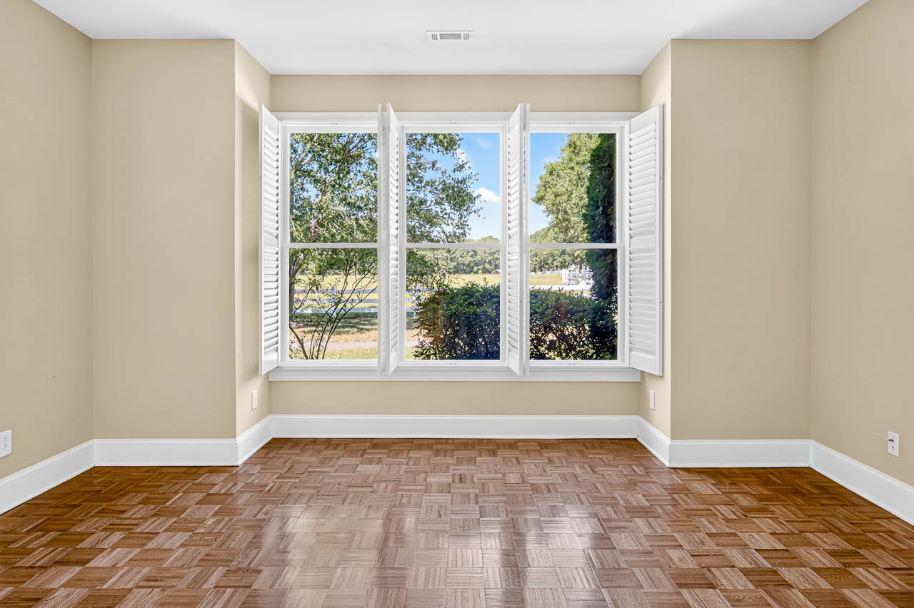 Empty room with beige walls, wood parquet floor, and a large window with white shutters open, revealing trees and greenery outside under a bright blue sky.