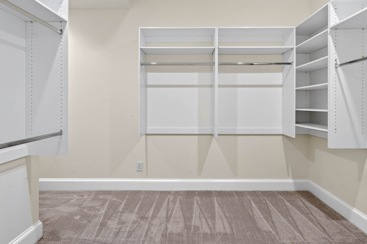 Empty walk-in closet with white shelving units, hanging rods, and beige carpet. The walls are light-colored, and the space appears clean and well-lit.