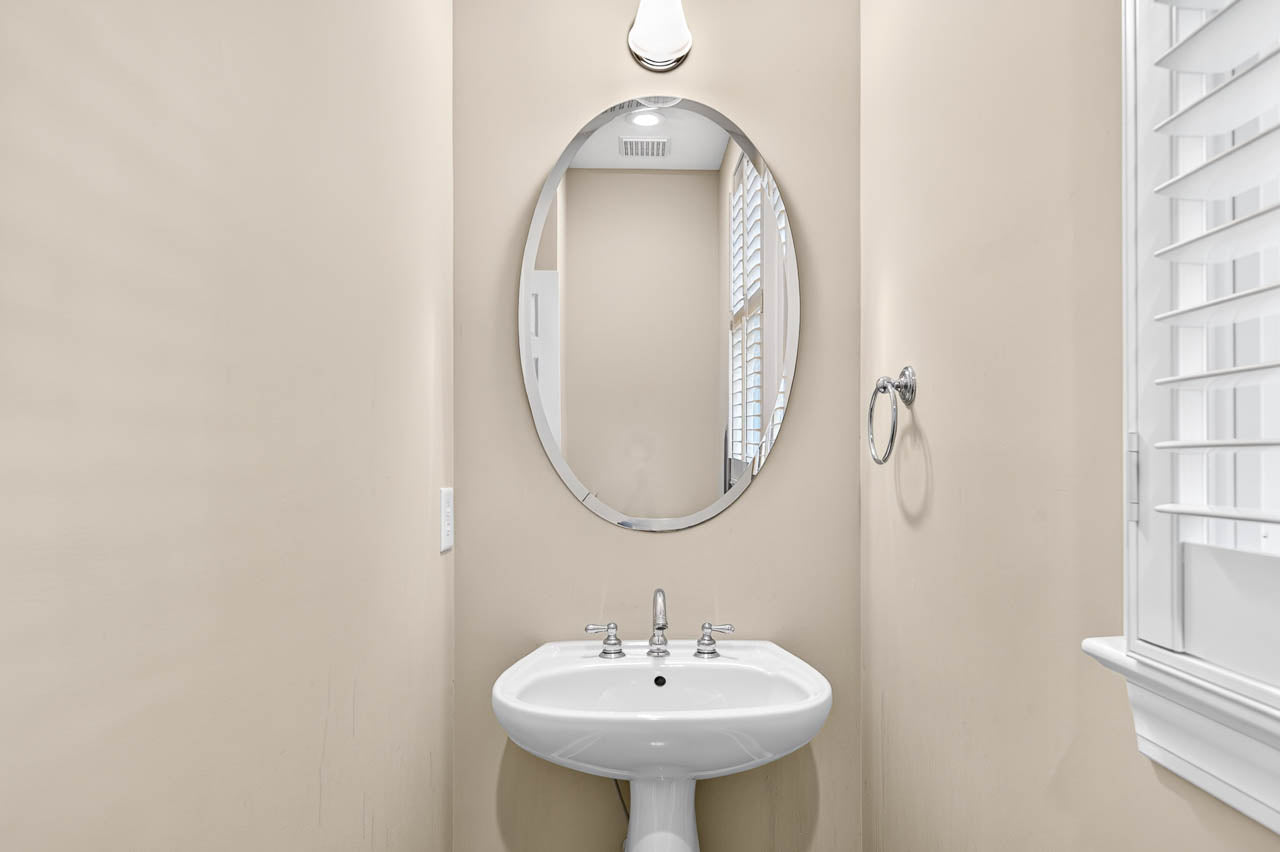 A small beige bathroom with an oval mirror above a white pedestal sink, a wall-mounted light, towel ring, and a window with white shutters on the right.