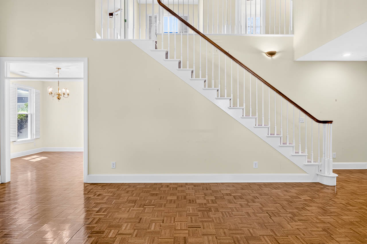 A staircase with white railings and wooden steps along a beige wall; to the left, an open doorway leads to a bright room with parquet flooring, windows, and a chandelier.