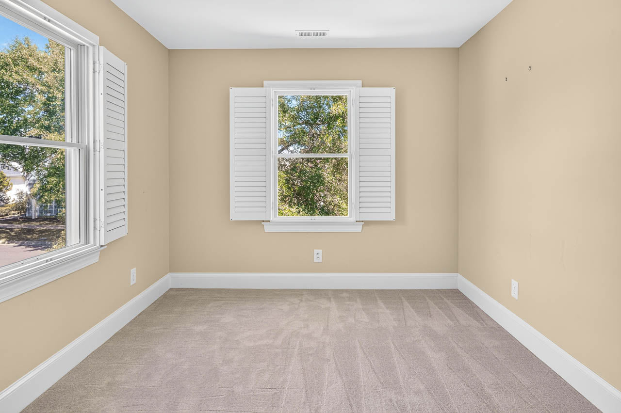 Bright empty room with beige walls, carpeted floor, and two windows with white shutters letting in natural light. Trees are visible through the windows.