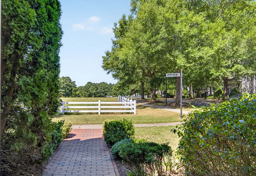 Brick path bordered by shrubs leads to a white fence and open grassy area, with large trees and a street sign visible to the right under a clear blue sky.