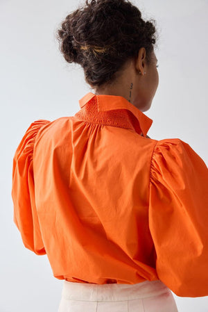 A person with curly hair is seen from behind wearing the HOPE FOR FLOWERS - SMOCKED SHIRT IN FLAME by HOPE FOR FLOWERS—an orange organic cotton poplin shirt with puff sleeves—and light pants. A numbered tattoo is visible on their neck against a plain background.
