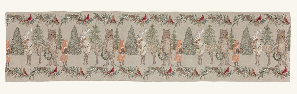 A CHRISTMAS CHEER EMBROIDERED LINEN TABLE RUNNER featuring woodland animals in winter attire and Christmas trees, draped over a wooden dining table set with plates, glassware, and candlesticks.