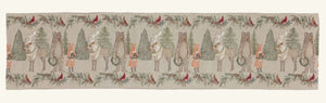 A CHRISTMAS CHEER EMBROIDERED LINEN TABLE RUNNER featuring woodland animals in winter attire and Christmas trees, draped over a wooden dining table set with plates, glassware, and candlesticks.