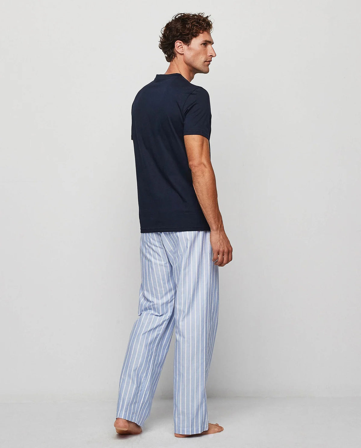 A man stands barefoot against a plain background, wearing the MIRTO 1956 - OXFORD PAJAMA SET by MIRTO 1956, featuring a dark blue top and light blue and white striped cotton pants with a comfortable drawstring waist.