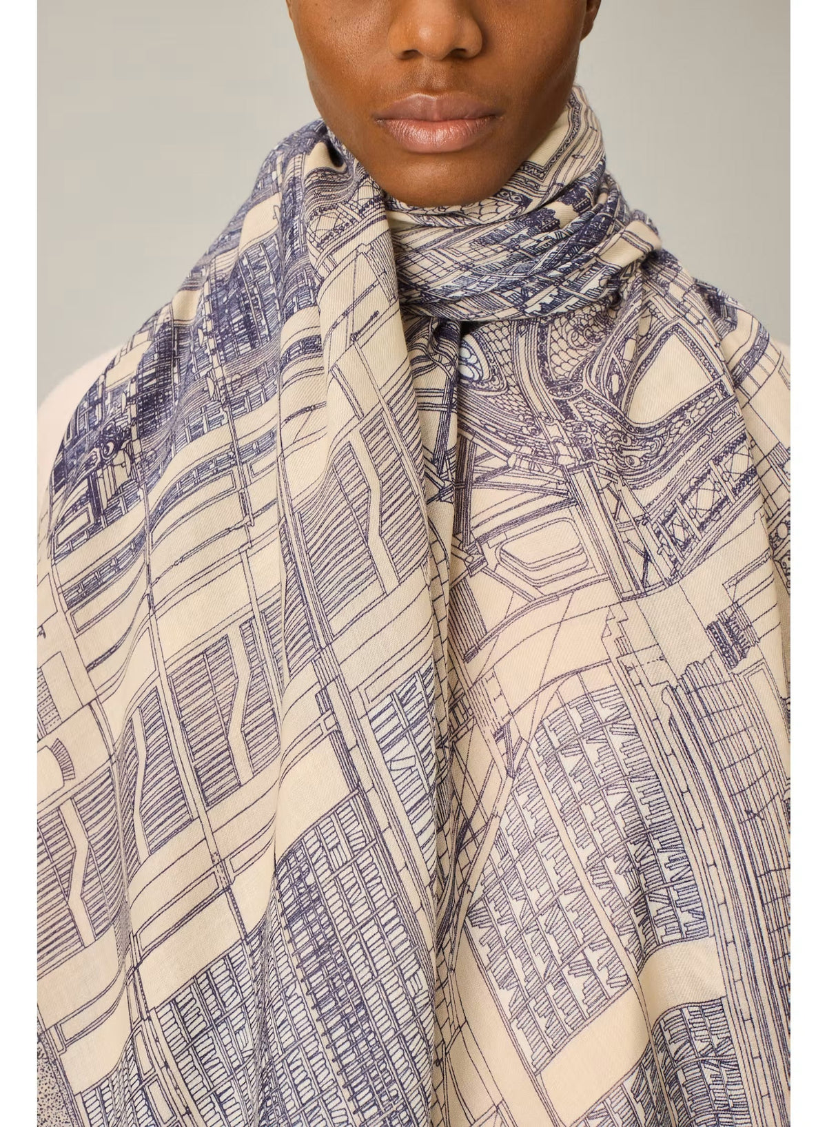 A person wearing a cream-colored outfit stands facing forward, draped in the INOUI EDITIONS VIVIENNE Wool Scarf in Grey—a large, patterned scarf featuring blue and beige tones. Made from 100% wool and crafted in India by INOUI, the scarf measures 100 x 190 cm and is set against a plain, light-colored background.
