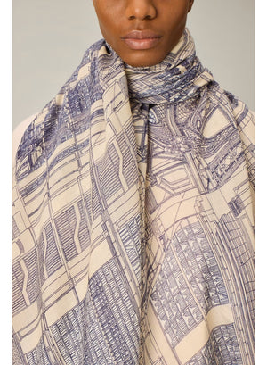 A person wearing a cream-colored outfit stands facing forward, draped in the INOUI EDITIONS VIVIENNE Wool Scarf in Grey—a large, patterned scarf featuring blue and beige tones. Made from 100% wool and crafted in India by INOUI, the scarf measures 100 x 190 cm and is set against a plain, light-colored background.
