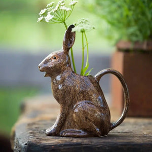 The QUAIL CERAMICS - HARE JUG, LARGE by QUAIL DESIGNS, a delightful ceramic pitcher shaped like a brown rabbit, sits on a wooden surface with white flowers artfully arranged in its open mouth. This charming decorative piece brings whimsy to the tabletop display, while green plants create a softly blurred background.