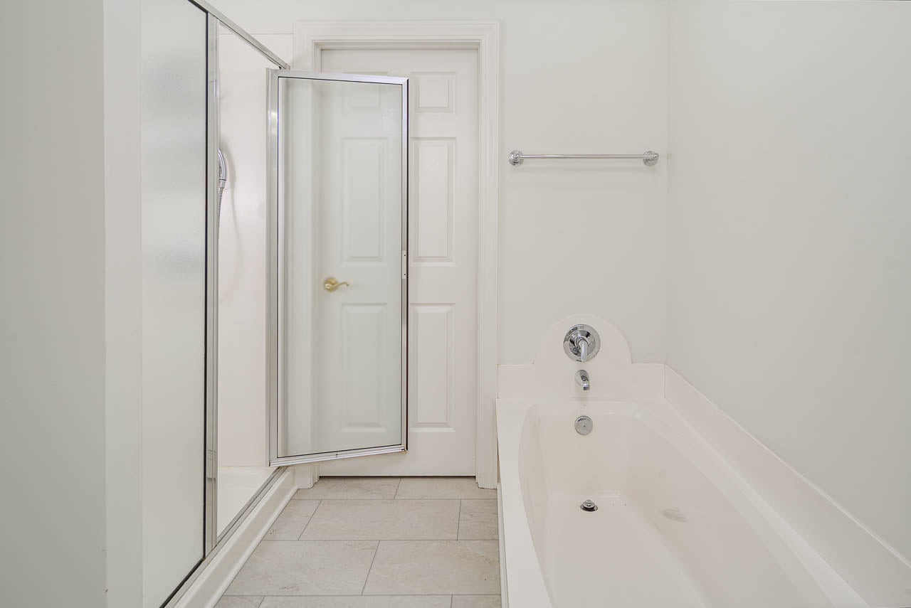 A clean, white bathroom with a built-in bathtub, a glass-enclosed shower, a towel bar on the wall, a tiled floor, and a closed door in the background.