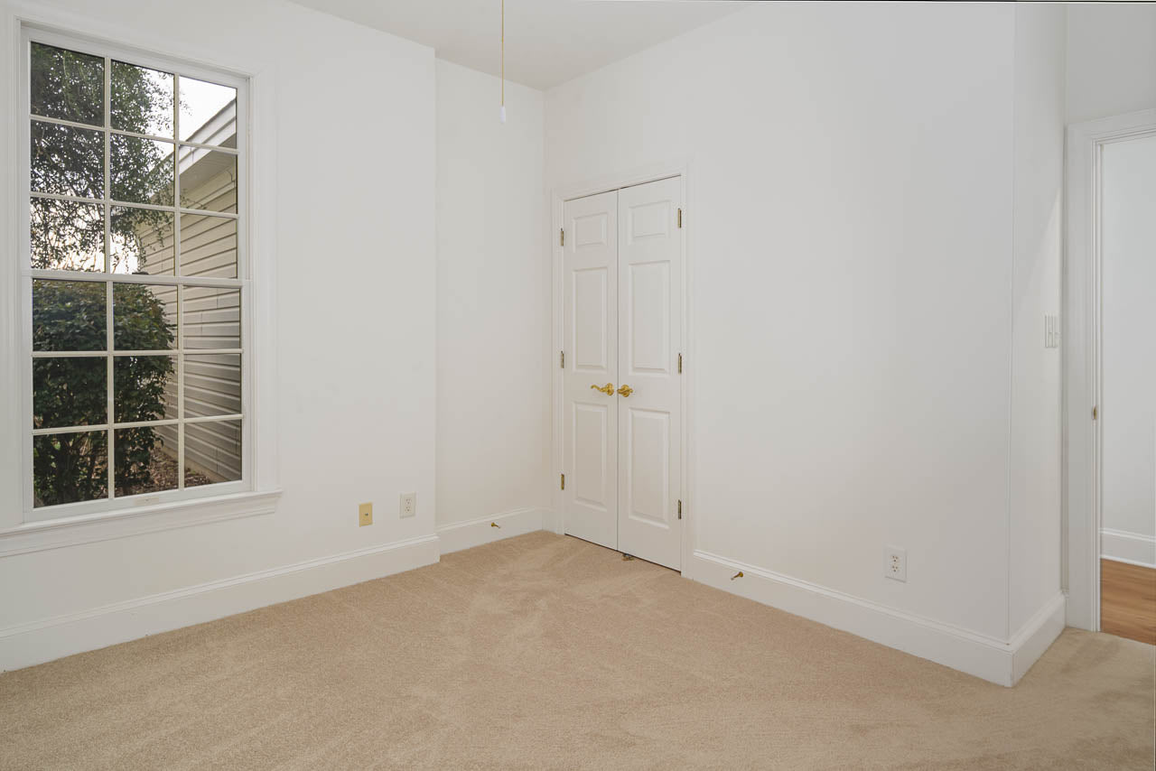 A small, empty room with beige carpet, white walls, a double-door closet, a large window showing greenery outside, and an open doorway leading to a hallway with hardwood floors.